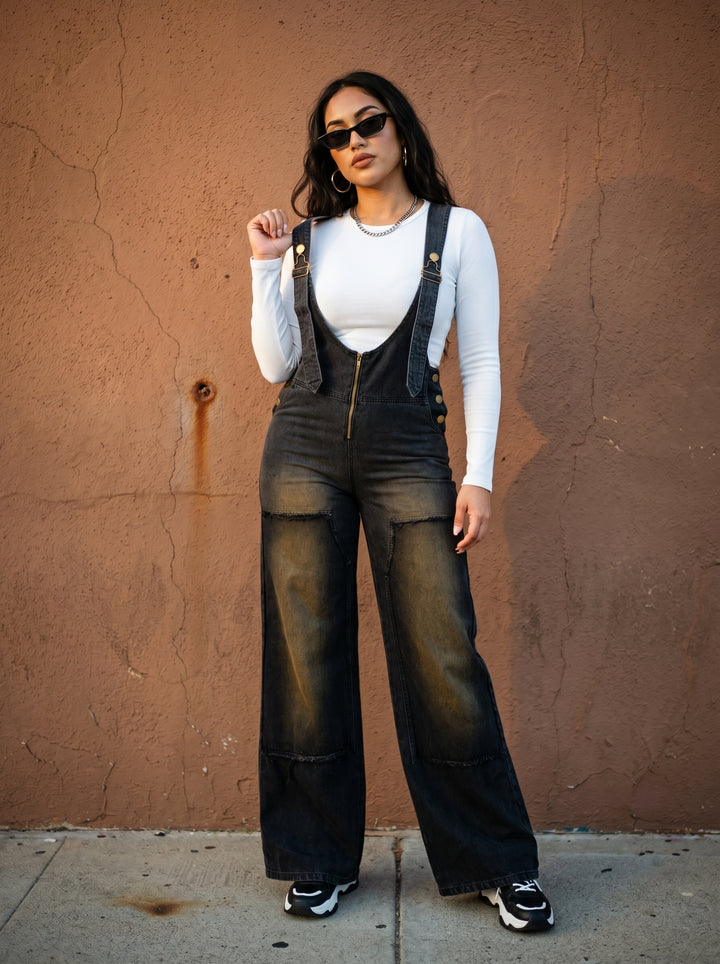 Woman wearing black overalls and sunglasses against a brown wall.