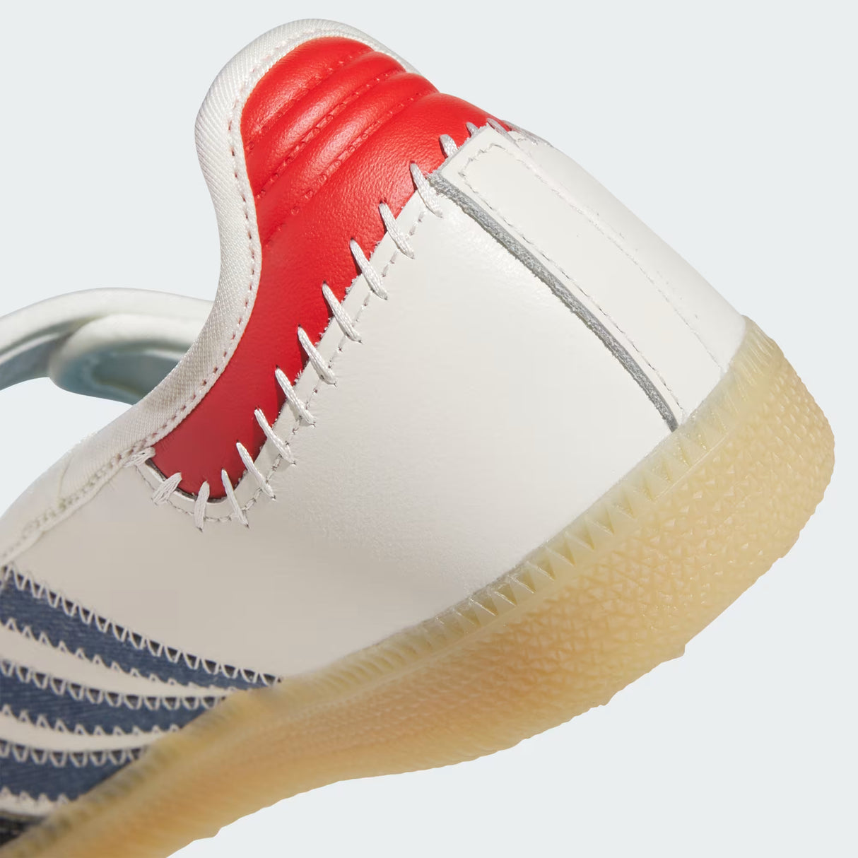Close-up of a white leather sneaker showing the heel and side profile, featuring a red heel counter, white contrast stitching, blue side stripes, and a translucent gum sole.