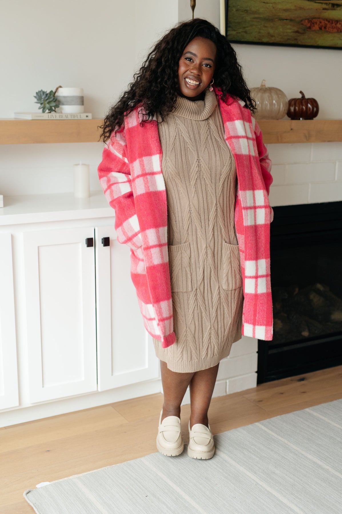 Woman wearing a beige sweater dress and red plaid coat in a home setting.