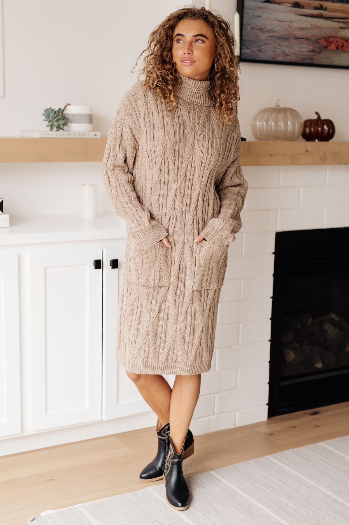Woman wearing a beige cable knit sweater dress standing in a room with a fireplace.