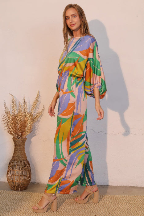 Woman wearing a colorful patterned jumpsuit standing against a plain wall with a woven plant vase to her left. side view