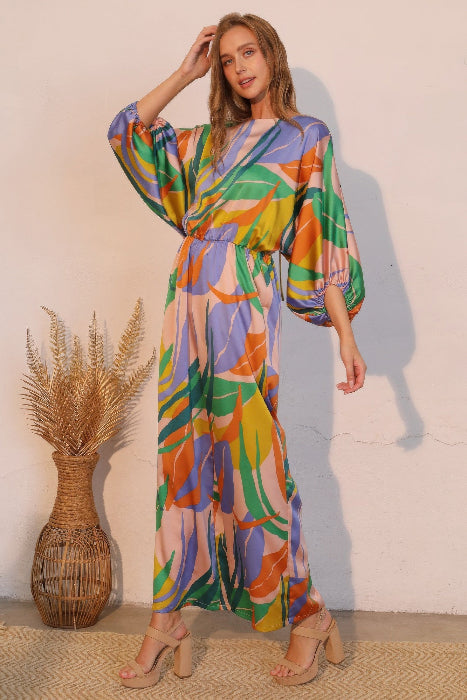 Woman wearing a colorful jumpsuit standing next to a woven vase against a plain wall.
