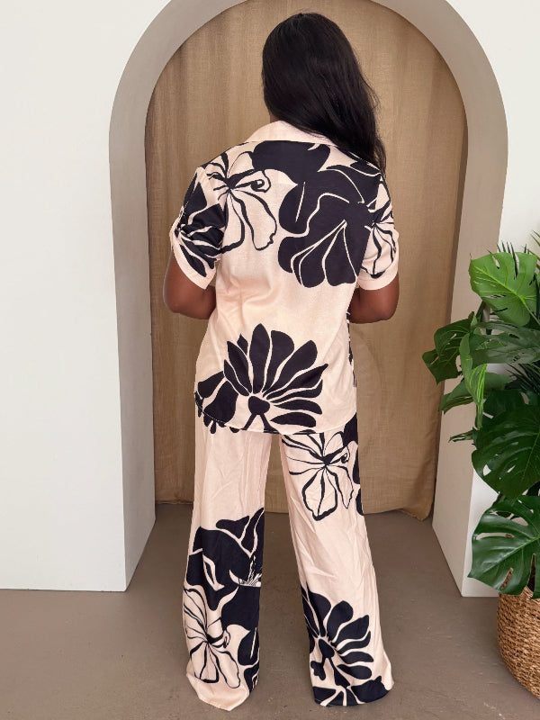 Woman wearing a floral pant set standing in front of a beige wall with a plant to the side.