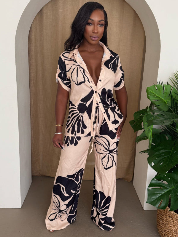 Woman wearing a beige and black floral pant set standing in front of a beige curtain with a plant to the side.