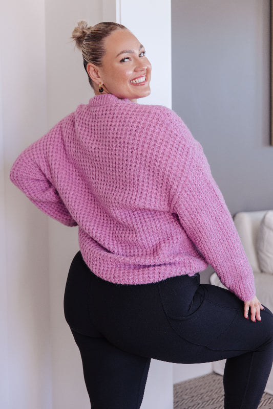 Woman wearing a pink knitted sweater and black pants sitting on a white chair.