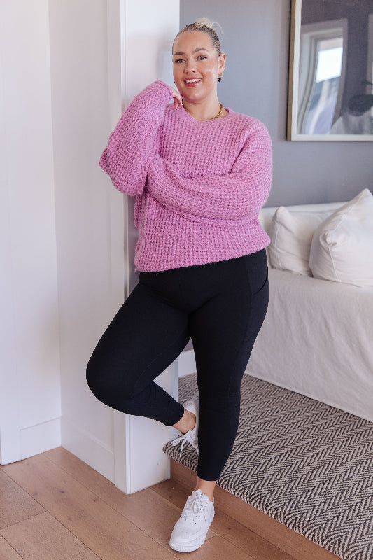Woman wearing a pink sweater and black leggings standing in a room with a couch and framed picture.