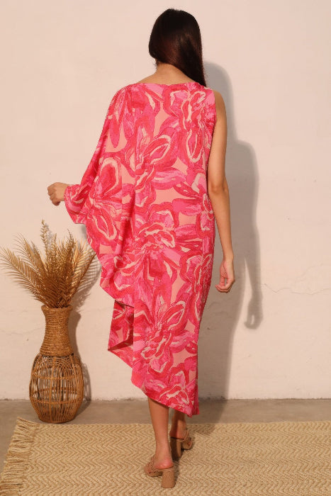 Woman wearing a pink floral dress showing the back view standing against a plain wall with a woven mat and vase on the floor.