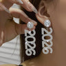 Close-up of a person wearing sparkling 2026 earrings with a hand holding one of them.