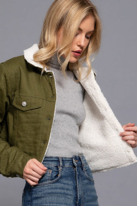 Woman wearing a green jacket with a white fleece lining, gray sweater, and blue jeans on a gray background