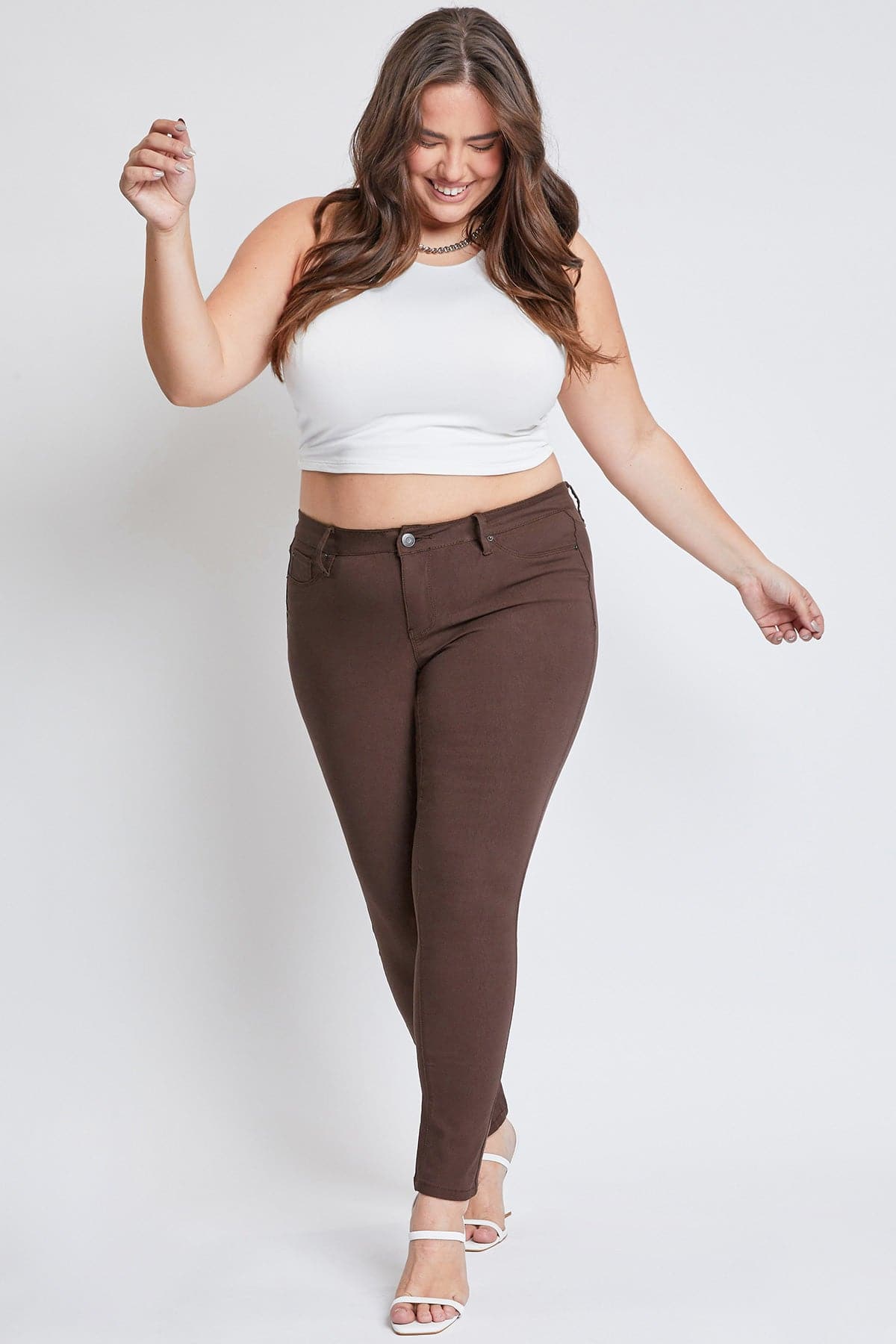 A woman modeling a white ribbed crop top and brown skinny jeans against a light gray studio background.