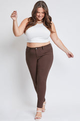 A woman modeling a white ribbed crop top and brown skinny jeans against a light gray studio background.