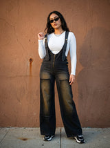 Woman wearing black overalls and sunglasses against a brown wall.