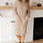 Woman wearing a beige cable knit sweater dress standing in a room with a fireplace.