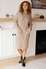 Woman wearing a beige cable knit sweater dress standing in a room with a fireplace.