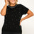 A woman modeling a black short-sleeve knit top with small white rhinestone-like dots scattered across the front and a rhinestone-trimmed crew neckline, paired with black pants against a plain light background.