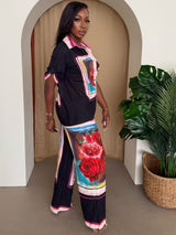 Woman wearing a black top with colorful design and floral pants standing in a room with a plant.