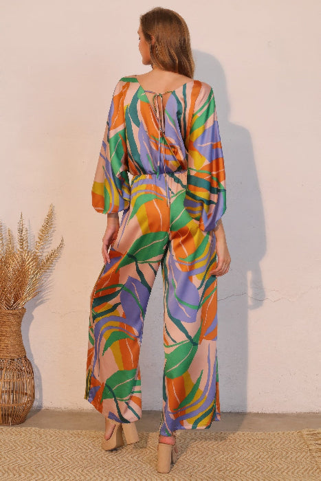 Woman wearing a colorful patterned jumpsuit standing against a plain wall. back view