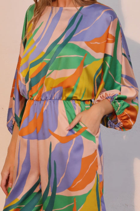 Close up of model with hand in her pocket wearing blue Colorful jumpsuit with a leaf pattern on a plain background