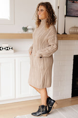 Woman wearing a beige cable knit sweater dress standing in a room with a fireplace.