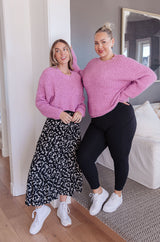 Two women wearing pink sweaters standing in a room with a mirror and bed in the background.