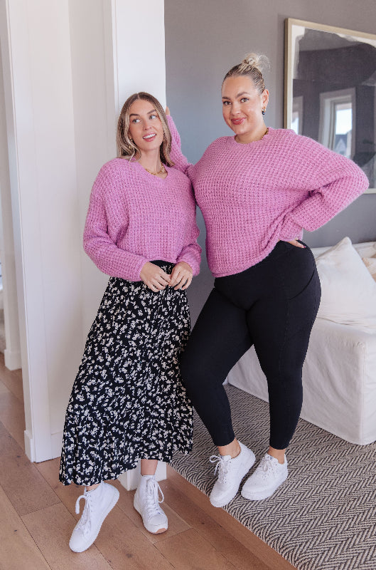 Two women wearing pink sweaters standing in a room with a mirror and bed in the background.