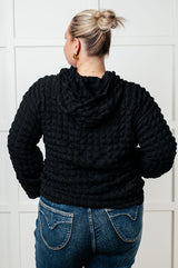 Person wearing a black textured hoodie and blue jeans against a white tiled wall.