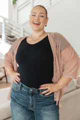 Woman wearing a black top and blue jeans with a pink cardigan indoors.