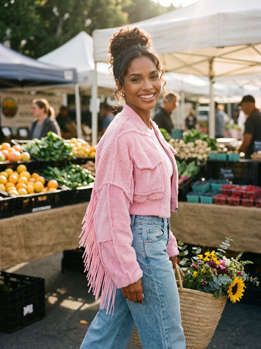 A woman at an outdoor farmers market wearing a light pink fringed suede jacket and light blue jeans, holding a basket of flowers.