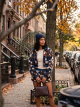 A woman stands on a city sidewalk wearing a brown, navy, and beige diamond-patterned zip-up jacket and matching shorts, a cream top, a navy beanie, and holding a brown handbag.