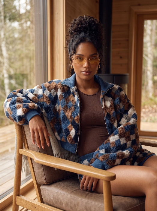 A woman wearing a blue, brown, and beige patterned sherpa zip-up jacket sits in a wooden chair in front of a rustic wood-paneled window.