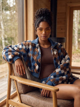 A woman wearing a blue, brown, and beige patterned sherpa zip-up jacket sits in a wooden chair in front of a rustic wood-paneled window.