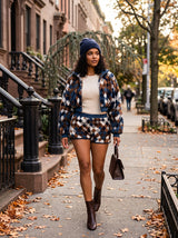 A woman walking on an autumn city sidewalk wearing a matching navy, brown, and beige argyle patterned jacket and shorts set, a cream top, a navy beanie, and brown ankle boots.