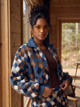 A woman wearing a blue, brown, and white diamond-patterned sherpa fleece zip-up jacket with a blue collar, standing on a wooden porch.
